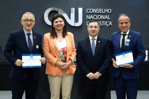 A imagem mostra quatro autoridades posando para foto em frente ao painel do CNJ. Da esquerda para a direita, estão o corregedor-geral da Justiça Militar do Estado de São Paulo, usando terno escuro e segurando um certificado; a conselheira do CNJ, vestindo blazer claro sobre blusa estampada; o secretário de Estratégia e Projetos do CNJ, de terno escuro e gravata; e o vice-presidente do TJMMG, também de terno, segurando um certificado. Todos estão em pé, lado a lado, em ambiente institucional.
