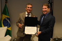 A imagem mostra dois homens usando terno. À esquerda está o ministro Artur Vidigal de Oliveira, que segura a comenda, e à direita, o desembargador Fernando Antônio Nogueira Galvão da Rocha, que entrega uma pasta com um diploma ao ministro. Ao fundo, no lado esquerdo, aparece uma bandeira do Brasil.