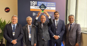 A imagem mostra cinco homens em pe, todos deterno. Eles estao em frente a um banner com a logomarca do 19° Encontro Nacional do Poder Judiciário, de cor azul. Da esquerda para a direita, estão o chefe de gabinete da Presidência do TJMMG, coronel Giovanne Gomes da Silva; o desembargador corregedor Sócrates Edgard dos Anjos; o desembargador vice-presidente Janmes Ferreira Santos, que segura o trofeu Excelência com os braços erguidos; o juiz de Direito do Juízo Militar Marcos Luiz Nery Filho; e o secretário especial da Presidência, Giovânni Viana Mendes 