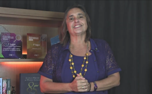 A imagem mostra a defensora publica Silvana LourençoLobo vestindo uma blusa azul e usando um colar amarelo. Ao fundo, ha uma prateleira com troféus da Justiça Militar e livros sobre Direito Militar. No canto direito da imagem está a janela de Libras, com a intérprete vestida de camiseta preta.