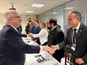 A imagem mostra o desembargador Janmes Ferreira Santos à direita, vestindo terno escuro, camisa branca e gravata marrom, enquanto cumprimenta o ministro Edson Faquin, à esquerda, que usa terno azul-marinho e gravata lilás. Ao fundo, outras autoridades também se cumprimentam em torno de uma mesa, em uma sala iluminada por luz branca e com paredes envidraçadas