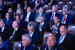 A imagem mostra os desembargadores do TJMMG sentados lado a lado na plateia durante a abertura do 19º Encontro Nacional do Poder Judiciário. Eles estão entre outras autoridades nas demais fileiras reservadas, todos estão vestidos de terno e gravata. As cadeiras são de cor marrom