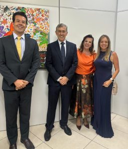 A imagem mostra quatro pessoas posando para foto em um ambiente interno. À esquerda, está o desembargador do TJMMG usando terno escuro e gravata amarela. Ao lado dele, está um homem usando terno azul-marinho e gravata escura. À direita, duas mulheres completam o grupo: uma delas veste blusa laranja e saia floral, enquanto a outra usa um vestido longo azul-marinho. Todos estão diante de uma parede clara com uma obra de arte colorida ao fundo.