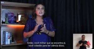 A imagem mostra a defensora pública Silvana Lourenço Lobo vestindo uma blusa azul e usando um colar amarelo. Ao fundo, há uma prateleira com troféus da Justiça Militar e livros sobre Direito Militar. No canto direito da imagem está a janela de Libras, com a intérprete vestida de camiseta preta.