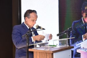 A imagem mostra o desembargador James Ferreira Santos  falando ao microfone enquanto segura uma folha de papel em uma tribuna. Ele veste um terno azul com gravata preta. À direita, um grande painel digital ao fundo exibe sua imagem ampliada.