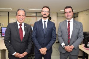 A imagem mostra três homens de terno, em pé. O primeiro à esquerda é o desembargador James Ferreira Santos, de terno preto, camisa branca e gravata vermelha.