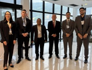 A imagem mostra seis participantes da 6ª Reunião Nacional de Gestão de Demandas do Eproc, sendo cinco homens e uma mulher, posando em um ambiente interno com amplas janelas de vidro. O segundo da direita para a esquerda da imagem é o juiz de Direito George Walter Barreto Paviótti, usando terno cinza escuro e gravata cinza.