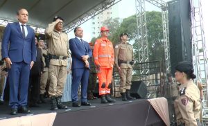 A imagem mostra cinco pessoas em pé sobre um palco, no pátio da Academia de Polícia Militar. O desembargador James Ferreira Santos é o primeiro da esquerda para a direita da imagem. Ele usa terno azul-escuro. As demais autoridades trajam terno e uniformes militares. À frente do palco está uma militar em posição de respeito