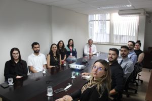 A imagem mostra um grupo de dez pessoas em uma sala de reuniões, sentados em torno de uma grande mesa de reunião, participando do programa Ambientar. Eles usam trajes formais. Há uma janela com cortina clara no fundo