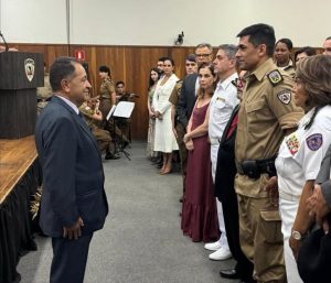 A imagem mostra uma fileira de pessoas em pé posicionadas de frente para um homem que usa roupa formal. Da direita para a esquerda, a terceira pessoa é o desembargador Jadir Silva. Ao fundo, aparecem militares fardados, músicos da corporação e convidados