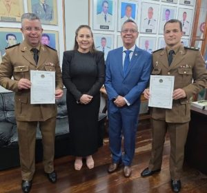 A imagem mostra quatro pessoas em pé, lado a lado, em um ambiente com paredes com quadros de autoridades ao fundo. À esquerda e à direita, dois oficiais fardados, seguram certificados com ambas as mãos. Ao centro, estão uma mulher vestindo traje preto, com as mãos entrelaçadas à frente do corpo, e um homem de terno azul, camisa clara e gravata, ambos sorrindo para a câmera. O grupo posa em uma sala com piso de madeira e iluminação interna