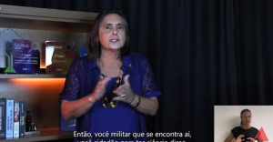 A imagem mostra a defensora pública Silvana Lourenço Lobo vestindo uma blusa azul e usando um colar amarelo. Ao fundo, há uma prateleira com troféus da Justiça Militar e livros sobre Direito Militar. No canto direito da imagem está a janela de Libras, com a intérprete vestida de camiseta preta.