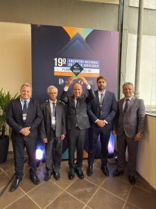 A imagem mostra cinco homens em pé, todos de terno. Eles estão em frente a um banner com a logomarca do 19º Encontro Nacional do Poder Judiciário, de cor azul. Da esquerda para a direita, estão o chefe de gabinete da Presidência do TJMMG, coronel Giovanne Gomes da Silva; o desembargador corregedor Sócrates Edgard dos Anjos; o desembargador vice-presidente Janmes Ferreira Santos, que segura o troféu Excelência com os braços erguidos; o juiz de Direito do Juízo Militar Marcos Luiz Nery Filho; e o secretário especial da Presidência, Giovânni Viana Mendes