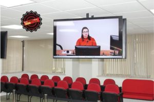 A imagem mostra uma sala de audiências do TJMMG. No teto está fixado um monitor de tela plana centralizado. A tela exibe a imagem da juíza Carolina Aleixo Benetti de Oliveira Rodrigues, uma mulher de cabelos escuros e óculos, vestindo uma blusa vermelha, sentada à frente de um computador em uma transmissão de vídeo. Abaixo do monitor estão poltronas de auditório com estofado vermelho. Ao fundo, janelas com persianas verticais em tom bege cobrem a parede. No canto superior esquerdo, uma câmera está instalada no teto, apontada para o auditório.