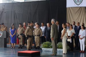 A imagem mostra autoridades civis e militares perfiladas, tendo ao centro o vice-presidente do TJMMG de terno escuro e camisa branca. O evento ocorre em um pátio coberto