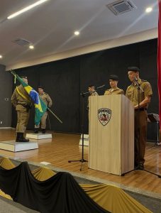 A imagem mostra cinco militares em um palco. O primeiro deles, à esquerda, segura a bandeira do Brasil. À direita, dois militares estão em pé atrás de uma tribuna de madeira com o brasão do PMMG, falando ao microfone. Ao fundo, outros dois militares permanecem perfilados no palco. O palco tem piso de madeira e fundo preto