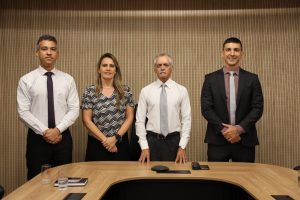 A imagem mostra quatro pessoas em pé à frente de uma mesa de reunião, sendo três homens e uma mulher. O primeiro à direita da imagem é o juiz Antônio Moreno Boregas e Rêgo, vestindo terno preto e gravata cinza. Ao seu lado está o desembargador Sócrates Edgard dos Anjos, com camisa branca, gravata cinza e calça escura. Eles estão na Sala Diamante do TJMMG, cujo ambiente tem paredes com revestimento em tons de madeira e preto, e uma mesa de reunião no mesmo tom.