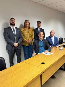 A imagem mostra cinco pessoas atrás de uma mesa em uma sala de reunião, sendo quatro homens e uma mulher. Três delas estão em pé e duas estão sentadas. A primeira pessoa sentada à esquerda é o juiz Antônio Moreno Boregas e Rêgo, de terno escuro e camisa azul. Ao seu lado está o advogado Cássio Antônio dos Santos, de terno azul e camisa clara. Em pé, da esquerda para a direita, está o promotor de Justiça Lucas Pena Forte Dias, de terno cinza, camisa branca e gravata listrada; a defensora pública Silvana Lobo, de calça jeans e camisa amarelo-mostarda; e o advogado Edmar Assis, de terno e gravata escuros e camisa branca