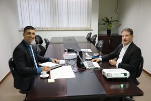 A imagem mostra dois homens sentados em uma mesa de reunião. À direita está o desembargador Fernando Antônio Nogueira Galvão da Rocha vestindo terno preto e camisa branca, e à esquerda está o juiz Antônio Moreno Boregas e Rêgo vestindo terno preto e camisa azul. Há uma janela com cortina clara no fundo.