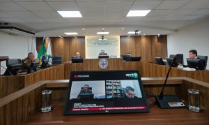 A imagem mostra os desembargadores durante a Sessão Ordinária no Tribunal Pleno, sentados em uma tribuna de madeira. Eles estão posicionados atrás de computadores e microfones. Em primeiro plano, um monitor exibe a transmissão da sessão. O ambiente apresenta paredes revestidas em madeira, bandeiras oficiais e o brasão do TJMMG ao fundo.
