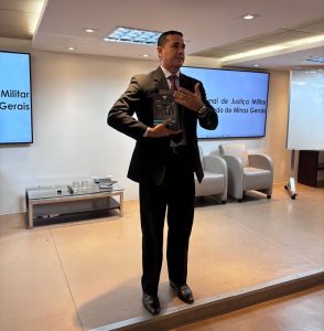A imagem mostra o coronel em pé, segurando um livro enquanto fala ao público no auditório do TJMMG. Ele está vestindo terno e gravata escuros. Ao fundo, há dois telões onde lê-se “Tribunal de Justiça Militar do Estado de Minas Gerais".