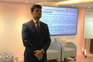 A imagem mostra o coronel em pé no auditório do TJMMG. Ele está vestindo terno escuro e gravata. Ao fundo, há um telão escrito “Competência da Justiça Militar” e um texto projetado.