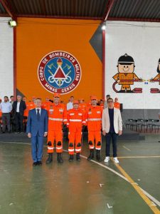 A imagem mostra cinco pessoas em pé, na sede do 2º Batalhão de Bombeiros Militar. A primeira pessoa à esquerda da imagem é o desembargador Osmar Duarte Marcelino, vestindo terno azul-escuro e gravata cinza. Ao lado está o tenente-coronel Herbert Aquino Marcelino, a coronel Amanda Cristina Miranda e o tenente-coronel Vitor Costa Leite, todos de uniforme laranja do Corpo de Bombeiros. À direita está o coronel veterano Luiz Henrique Gualberto Moreira, de camisa e calça escuras, e blazer bege. Ao fundo, há o dispositivo de honra com outras autoridades civis e militares, e na parede um grande painel laranja com o brasão da corporação escrito “Corpo de Bombeiros Militar de Minas Gerais”.
