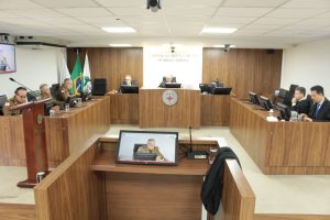 A imagem mostra os desembargadores durante a Sessão Ordinária no Tribunal Pleno, sentados em uma tribuna de madeira. Eles estão posicionados atrás de microfones e computadores. O ambiente apresenta paredes revestidas em madeira, bandeiras oficiais e o brasão do TJMMG ao fundo. 