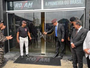 A imagem mostra um grupo de aproximadamente cinco pessoas, entre militares fardados e autoridades, reunido em frente à entrada do memorial ROTAM. Na porta de vidro há um laço preto fixado. À direita da imagem, o desembargador Rúbio Paulino Coelho, vestido com terno escuro, aparece com a mão estendida em direção à porta, enquanto os demais presentes observam a ação.