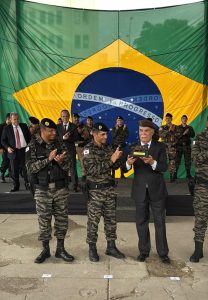 A imagem mostra três homens em primeiro plano durante a solenidade de 45 anos da ROTAM. Da direita para a esquerda, o primeiro é o desembargador Rúbio Paulino Coelho que veste um terno preto e gravata azul, e segura um prêmio. Atrás aparecem diversos homens, entre eles, o desembargador James Ferreira Santos, o coronel Giovanne Gomes da Silva e militares. Ao fundo, aparece uma enorme bandeira do Brasil.