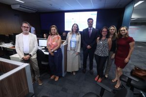 A imagem mostra sete pessoas em pé, entre homens e mulheres, uma ao lado da outra, na sala Lapidar do TJMG. No centro, está o desembargador Fernando José Armando Ribeiro, vestindo um terno cinza e gravata roxa.Todos estão usando roupas sociais e casuais. Ao fundo, aparece um grande painel de telas exibindo gráficos.
