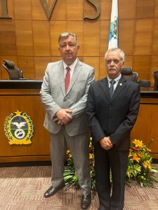 A imagem mostra dois homens em pé, no Plenário Ministro Waldemar Zveiter. À direita está o desembargador Sócrates Edgard dos Anjos, vestindo terno preto e gravata cinza. À esquerda está o desembargador Cláudio Brandão de Oliveira, vestindo terno cinza e gravata vermelha. Ao fundo, há uma parede revestida em madeira, uma mesa e uma bandeira.