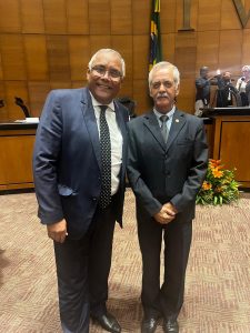 A imagem mostra dois homens em pé, no Plenário Ministro Waldemar Zveiter, com paredes revestidas em madeira. À direita está o desembargador Sócrates Edgard dos Anjos, vestindo terno preto e gravata cinza. À esquerda está o desembargador Gilberto Barbosa Batista dos Santos, vestindo terno azul escuro e gravata preta com bolinhas brancas. Ao fundo, há uma parede revestida em madeira, uma mesa e uma bandeira.