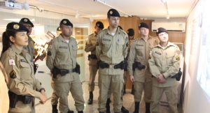 A imagem mostra um grupo de alunos do Curso de Formação de Soldados no Memorial da Justiça Militar. Eles estão uniformizados, com fardas bege e boinas pretas, observando o conteúdo exposto em uma das paredes do espaço.
