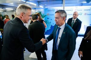 A imagem mostra dois homens em pé, cumprimentando-se com um aperto de mãos. À direita está o desembargador Osmar Duarte Marcelino, vestindo terno azul escuro e gravata azul. À esquerda está o desembargador Heráclito Vieira de Sousa Neto, vestindo terno preto. Eles estão em um ambiente com outras pessoas conversando