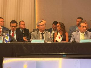 A imagem mostra os representantes do TJMMG e do TJMG na mesa de abertura do Consepre. Eles estão sentados lado a lado. Ao centro está o coronel Osmar Duarte Marcelino, de cabelos grisalhos, óculos, terno cinza, camisa branca e gravata escura. À direita da imagem está o presidente do TJMG, que usa terno claro com gravata verde. Ao fundo há vários homens e mulheres. Entre eles, está o chefe de gabinete da Presidência do TJMMG, de terno escuro, gravata escura e camisa azul