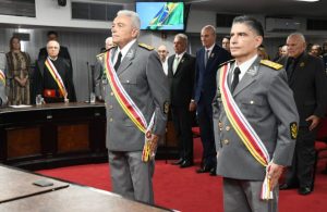 A imagem mostra dois homens em trajes militares formais, posicionados lado a lado em primeiro plano, em ambiente interno do plenário do STM. Ambos vestem uniformes cinza, de generais do Exército Brasileiro, e carregam faixa diagonal multicolorida sobre o peito, composta por listras em vermelho, amarelo e branco, utilizada pelos ministros do STM. Os dois estão em posição de sentido, em atitude de respeito. Ao fundo, sobre um carpete vermelho, estão outras autoridades, entre homens e mulheres em pé, com trajes formais.