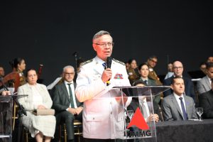 A imagem mostra o momento do discurso de posse do desembargador Osmar Duarte Marcelino, ele vestido com farda branca do Corpo de Bombeiros Militar de Minas Gerais. Ele está de pé, atrás de uma tribuna de acrílico com a logomarca do TJMMG, e segura um mcrofone. Atrás estão sentadas diversas autoridades, entre homens e mulheres vestindo trajes formais. Ao fundo estão músicos da Orquestra Sinfônica da Polícia Militar. 