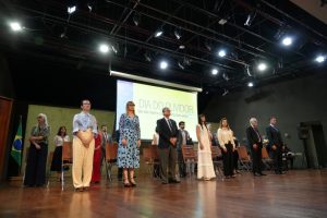 A imagem mostra três fileiras de pessoas, entre homens e mulheres, no palco do auditório JK. Na primeira fila, à esquerda da imagem, está a juíza Daniela de Freitas Marques, vestindo blusa azul e calça bege. A quarta pessoa da fila é a ouvidora-geral Gabriela Câmara Campos Bernardes Siqueira, usando um vestido longo branco. Ao fundo, há uma tela de projeção escrita: Dia do Ouvidor. Um dia inteiro de capacitações para você