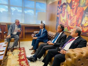 A imagem mostra quatro homens sentados em um gabinete do TRT3. Todos vestem ternos e usam gravatas. O primeiro à direita da imagem é o desembargador Sebastião Geraldo de Oliveira, sentado em uma cadeira, segurando um convite em mãos. Ele veste terno e gravata cinza, camisa branca, e está voltado para o desembargador Osmar Duarte Marcelino, que está sentado na ponta de um sofá, de terno e gravata azul, e camisa branca. Ao seu lado, no sofá, está o desembargador James Ferreira Santos, de terno escuro, camisa branca e gravata azul, e o coronel Giovanne Gomes da Silva, de terno escuro, camisa branca e gravata vermelha. Na parede à direita, há um quadro grande e colorido, e ao fundo da sala há uma ampla janela acima de um aparador de madeira.
