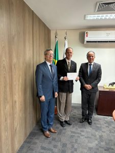 A imagem mostra três homens em pé, um ao lado do outro, em uma sala com a parede amadeirada. O primeiro à esquerda é o desembargador Osmar Duarte Marcelino, vestindo terno e gravata azul, e camisa branca; o segundo é o desembargador Júlio César Lorens, que está segurando um convite e vestindo paletó preto e calça clara; e o terceiro é o desembargador James Ferreira Santos, vestindo terno preto e gravata azul com camisa branca. Ao fundo, aparecem duas bandeiras.