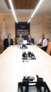 A imagem mostra quatro homens sentados ao redor de uma mesa de reunião. O primeiro à esquerda da imagem é o desembargador James Ferreira Santos, vestindo terno preto, camisa branca e gravata azul; o segundo é o presidente da OAB-MG, José Alberto Ribeiro Simonetti, vestindo terno cinza, camisa branca e gravata azul, segurando o convite juntamente com o desembargador Osmar Duarte Marcelino, que é o terceiro vestindo terno e gravata azul, e camisa branca. O quarto é o desembargador Rúbio Paulino Coelho, vestindo camisa branca e gravata roxa, e com o braço direito em uma tipoia. Ao fundo, há um painel na parede com a logomarca da OAB-MG escrito “Unir para avançar, forte e presente”. À esquerda da imagem aparece uma bandeira.