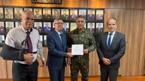 A imagem mostra quatro homens em pé, um ao lado do outro, em um ambiente institucional com paredes revestidas em madeira, tendo ao fundo um painel escrito “comandantes” e uma galeria de retratos. O primeiro à esquerda da imagem é o desembargador Rúbio Paulino Coelho, de camisa clara, gravata bordô e calça escura, com o braço direito em uma tipoia. O segundo é o desembargador Osmar Duarte Marcelino, de terno e gravata azul, e camisa clara. Ele segura o convite da posse da nova gestão do TJMMG juntamente com o general de Divisão Adriano Fructuoso da Costa, vestindo uniforme camuflado do Exército. O último à direita da imagem é o desembargador James Ferreira Santos, de terno cinza escuro, camisa clara e gravata azul.