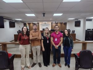 A imagem mostra cinco pessoas em pé, uma ao lado da outra, no Plenário do TJMMG. O grupo é formado por três mulheres e dois homens, todos trajando roupas sociais ou no estilo casual elegante. A primeira pessoa à direita da imagem é a servidora Cynthia Chiari, que veste blusa vermelha e calça clara; a segunda é Leonardo Carlotti Passos, usando camisa marrom e calça clara; a terceira é Rosângela Queiroz de Mello, com blusa e calça pretas; o quarto é Bruno Mendonça Cipriano, trajando camisa rosa e calça escura; e a quinta pessoa, Bárbara Marques, que veste blusa preta e calça azul. Eles estão em pé, diante de uma tribuna de madeira, tendo ao fundo uma parede branca escrita Tribunal de Justiça Militar do Estado de Minas Gerais