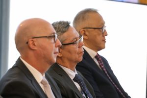 A imagem mostra três homens sentados um ao lado do outro, vistos de perfil e voltados para a direita. Ao centro está o desembargador Osmar Duarte Marcelino, vestindo terno preto e gravata azul-escura; à esquerda está o desembargador Rodrigo Mohr Picôn, usando terno preto e gravata clara; e, à direita, o desembargador Silvio Hiroshi Oyama, trajando terno preto e gravata preta com detalhes em vermelho. O fundo da imagem é claro e desfocado, destacando os três homens em primeiro plano.