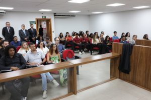 A imagem mostra os alunos da Faculdade de Direito da Fumec durante sessão no Tribunal Pleno. Eles estão sentados em cadeiras vermelhas do Plenário do TJMMG. As paredes ao fundo são brancas e há uma bancada madeira em primeiro plano
