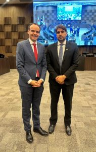 A imagem mostra dois homens em pé, um ao lado do outro, no auditório do Tribunal Pleno do TJMG. À direita da imagem, está o juiz Marcos Luiz Nery Filho, trajando terno preto, camisa branca e gravata cinza. À esquerda, está o desembargador Edilson Vitorelli, vestindo terno azul escuro, camisa branca e gravata vermelha. Ambos estão com as mãos à frente do corpo. Ao fundo, há um grande telão que exibe a imagem do plenário.