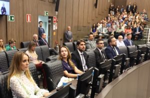 A imagem mostra o auditório do Tribunal Pleno do TJMG, com várias pessoas sentadas em fileiras. Em primeiro plano, há uma mesa longa com computadores e microfones, onde estão homens e mulheres vestidos formalmente. Entre eles, a terceira pessoa ao centro da imagem é o juiz Marcos Luiz Nery Filho, usando terno preto e gravata cinza. Ao fundo, o auditório está com o público acomodado em assentos em formato de arquibancada. As paredes do ambiente são revestidas em madeira.