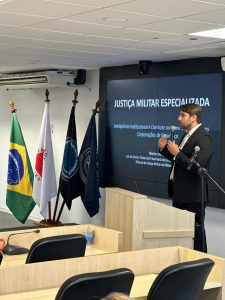 : A imagem mostra o juiz Marcos Luiz Nery Filho em pé, vestindo terno preto, segurando um microfone, durante uma palestra. Ao fundo, há uma tela onde está escrito “Justiça Militar Especializada. Inteligência Institucional e Combate ao Crime Organizado nas Corporações de Segurança”. À esquerda, estão quatro bandeiras. 