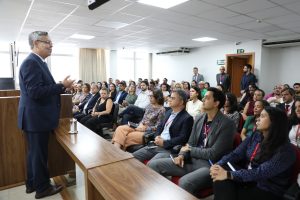 A imagem mostra o desembargador Osmar Duarte Marcelino usando terno azul escuro, em pé, atrás de uma bancada de madeira, na sala do Tribunal Pleno. À sua frente, há várias pessoas, entre homens e mulheres, sentadas em cadeiras, vestindo roupas formais.